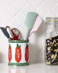 tomato can holding kitchen tool items on a kitchen counter with 2 white containers with wooden lids and a glass of back and white peas next to it. and a tiled backdrop