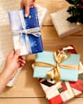 Overhead shot of someone handing over a gift wrapped in silver and blue wrapping paper, and tied in a silver bow, over a light wood surface.  On the light wood surface theres an assortment of other wrapped gifts and gift bags.