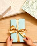 Overhead shot of two hands holding a small gift wrapped in green, white and gold wrapping paper.  In the top of the shot there are 3 other gifts wrapped in assorted white and gold paper.