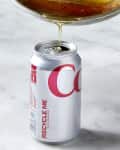 Angled shot of bacon grease being poured into a diet coke can on a white surface.
