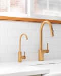 Gold kitchen faucets with white subway tile backsplash and a potted plant on the counter.