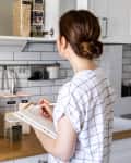 Woman in a kitchen writing in a notebook, surrounded by labeled food containers and kitchen utensils.