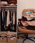 Wardrobe filled with hanging clothes, a chair piled with folded garments, and two woven baskets on the floor.