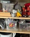 Cluttered cabinet with plastic bins, candles, baking tools, and kitchen items on wooden shelves.