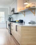 Modern kitchen with light wood cabinets, stainless steel appliances, and open shelving with jars and a blue toaster.