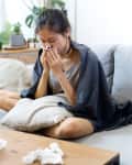 Sick woman sneezing at home on the sofa with a cold, she is covering with a blanket and blowing her nose