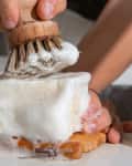 Hand scrubbing a foamy soap block with a wooden brush on a soap dish.
