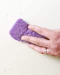 Hand cleaning a surface with a purple sponge, removing water droplets.