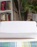 White wedge pillow on a bed with a colorful striped blanket, next to a bookshelf and potted plant.