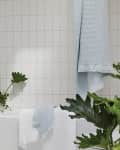 Bathroom with white tiles, pastel towels hanging, and green plants around a bathtub.