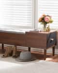 Wooden storage bench with gray cushion, books, flowers, and a gold vase. Tan heeled sandals and a gray hat on the floor.