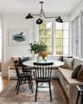 Dining nook with round white table, black chairs, built-in bench, and large windows. Vase with flowers on table.