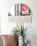 Half-circle mirror with fringe, potted plant, and textured cushion on a chair in a bright room.