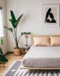 Minimalist bedroom with a wooden bed, gray bedding, abstract art, and potted plants on a patterned rug.