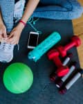 Person tying shoelaces on exercise mat with dumbbells, green ball, water bottle, and smartphone nearby.