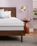 Modern bedroom with a wooden bed frame, white mattress, assorted pillows, and a gold lamp on a wooden side table.
