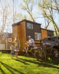 Family enjoying a sunny day outside tiny houses, with a picnic table, parked truck, and bicycle on green grass.