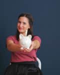 Woman in a pink shirt holding a white piggy bank against a dark blue background.