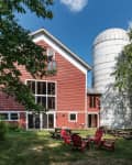 Red barn with large windows, adjacent white silo, surrounded by trees, and red Adirondack chairs on grass.