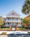Pink two-story house with white columns and balconies, flanked by green and red brick homes, palm tree in foreground.
