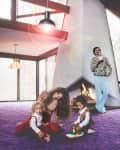 Family in a retro living room with purple carpet, large windows, and a geometric fireplace.