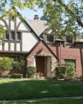 Tudor-style brick house with gabled roof, surrounded by trees and shrubs.