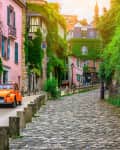 Orange vintage car driving down a cobblestone street lined with ivy-covered buildings and pink facades.