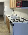 Compact kitchen with wooden table and chairs, gas stove, microwave, and a bed in the background.