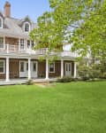 Three-story house with shingle siding, white columns, wraparound porch, and lush green lawn surrounded by trees.