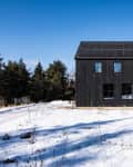 Black modern house with solar panels, surrounded by snow and trees under a clear blue sky.