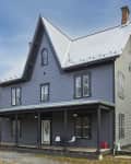 Gray two-story house with a steep metal roof, front porch, and surrounding trees.