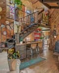 Rustic living room with wood paneling, staircase, bar stools, wood stove, and large windows with lace curtains.
