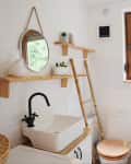 Bathroom with wooden shelves, round mirror, white sink, black faucet, and window view of greenery.