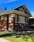 Two-story brick house with a front porch, large windows, and landscaped yard, under a clear blue sky.