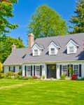 Traditional white house with gray roof, black shutters, and manicured lawn, surrounded by vibrant trees and shrubs.