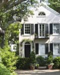 Two-story house with black shutters, arched windows, and lush greenery in the front yard.