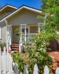 Cozy beige bungalow with white trim, front porch, and picket fence, surrounded by lush greenery and blooming flowers.