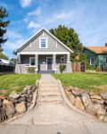 Gray house with white trim, front porch, and stone-lined walkway, surrounded by green lawn and trees.