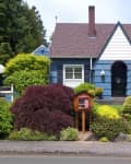 Blue cottage with brown roof, white trim, and lush garden featuring various shrubs and a small wooden library box.