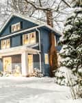 Blue two-story house with white trim, snow-covered yard, picket fence, and large tree in winter setting.