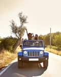 Blue Jeep with people enjoying a sunny drive on a rural road, surrounded by trees and grass.