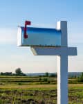 Metal mailbox with red flag on a white post, set against a rural landscape with fields and trees.