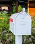 A white mailbox with a red flag in front of lush green plants and a wooden structure.