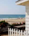 Beachfront cottage with white picket fence, red flowers, and ocean view. People walking on sandy beach in the background.