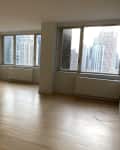 Empty room with light wood floors, large windows, and cityscape view. Red "Before" banner in the top left corner.