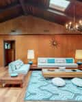 Mid-century modern living room with vaulted ceiling, skylights, white sofa, turquoise accents, geometric rug, and chandeliers.