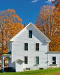 White farmhouse surrounded by vibrant autumn trees with orange leaves under a clear blue sky.
