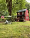 Red two-story cabin with a porch, surrounded by trees, picnic table with umbrella, and a wooden sauna in the yard.