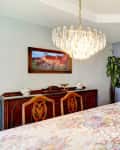 Dining room with a floral tablecloth, wooden sideboard, chandelier, and a view into a cozy living room.