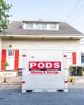 Moving and storage container in front of a beige house with red shutters, parked bicycle, and one-way street sign.